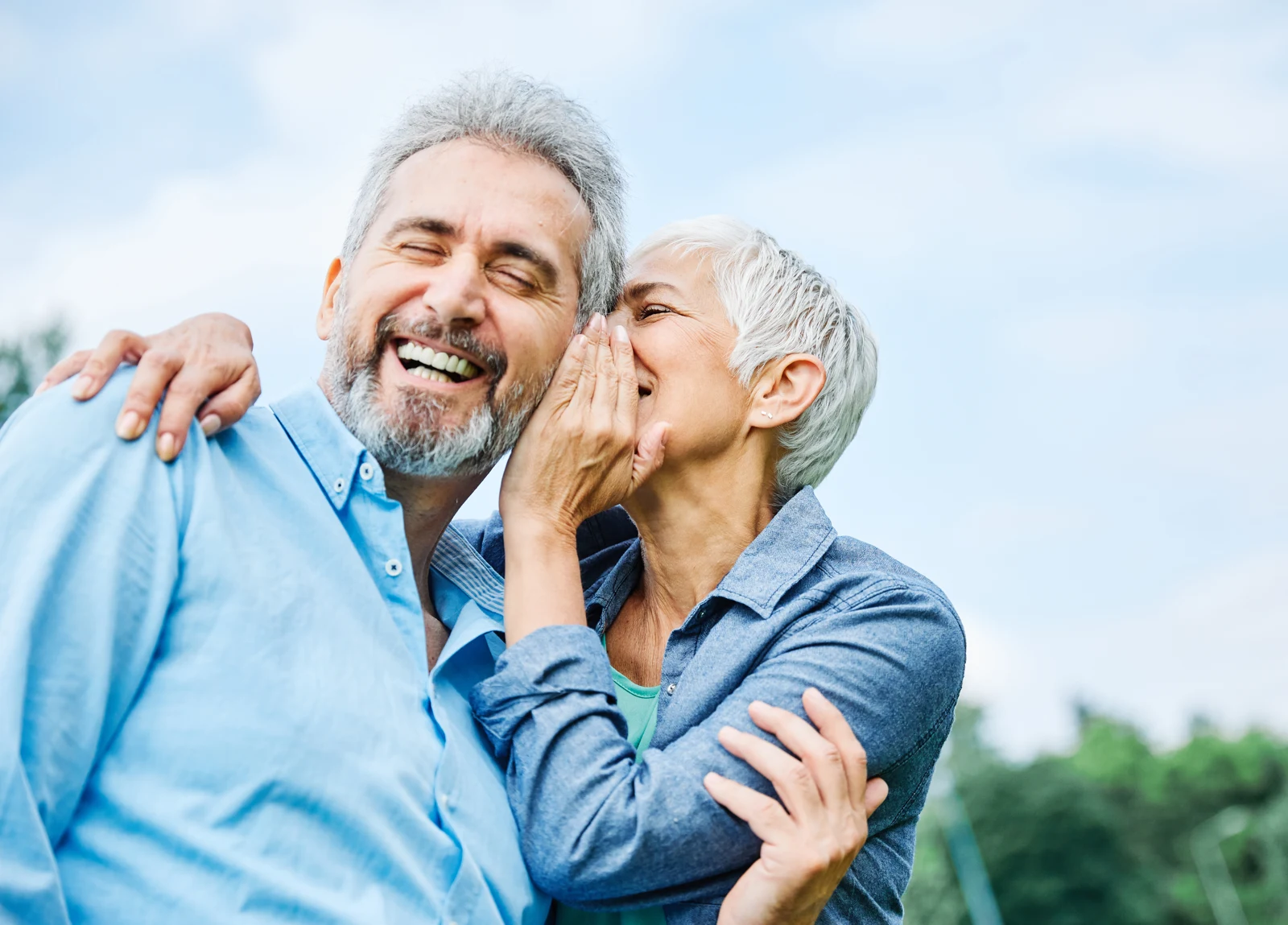 An older couple outdoors, one leaning close to the other’s cheek as they laugh.