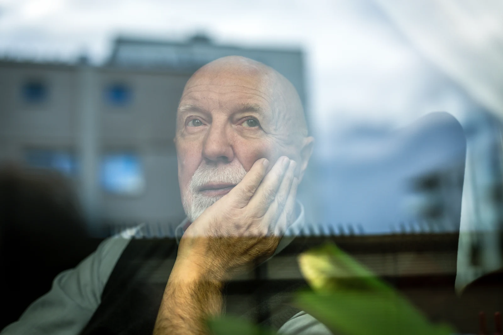 An older man resting his face on his hand beside a reflective window.