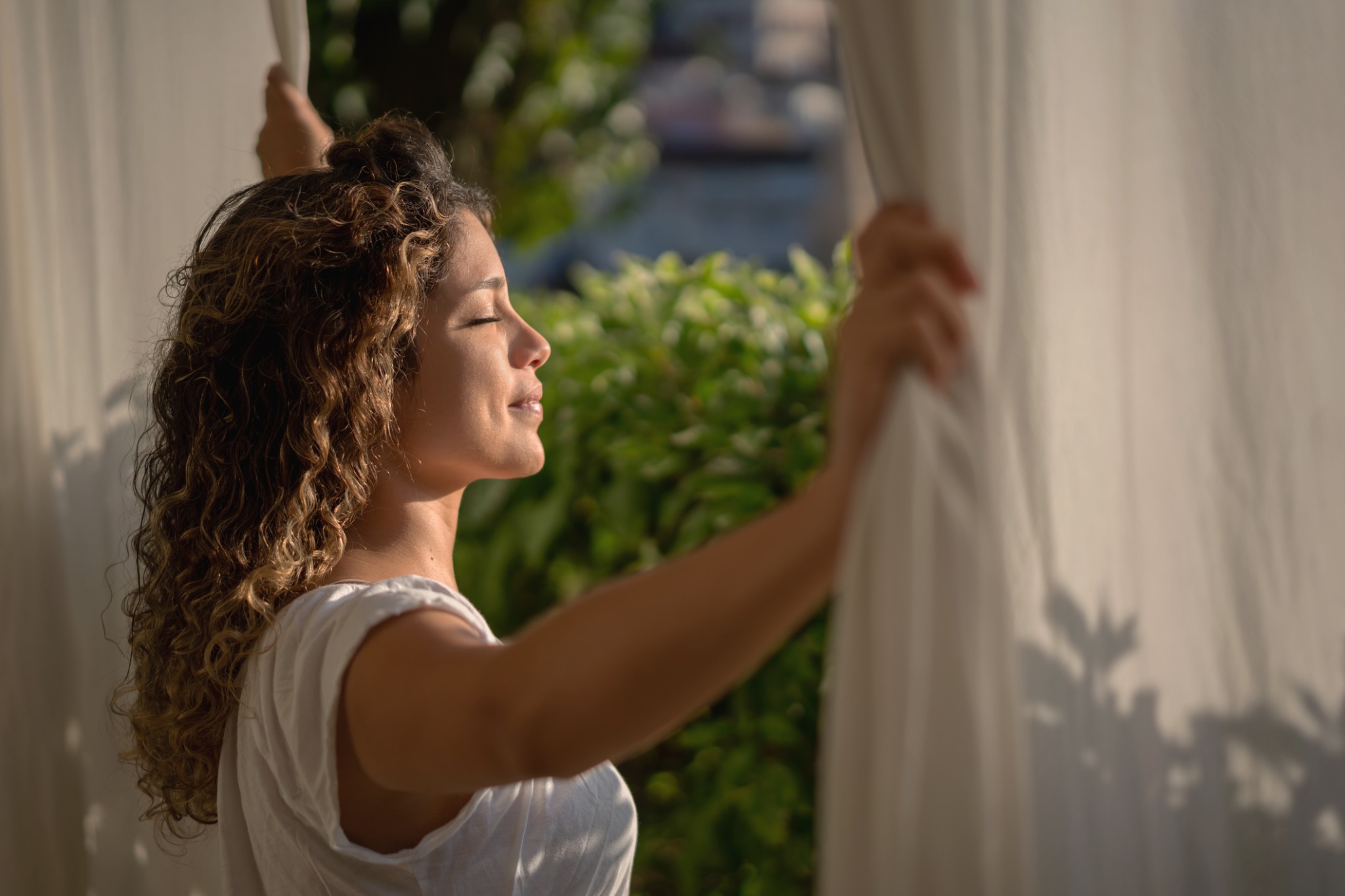 A woman opening curtains with sunlight on her face.
