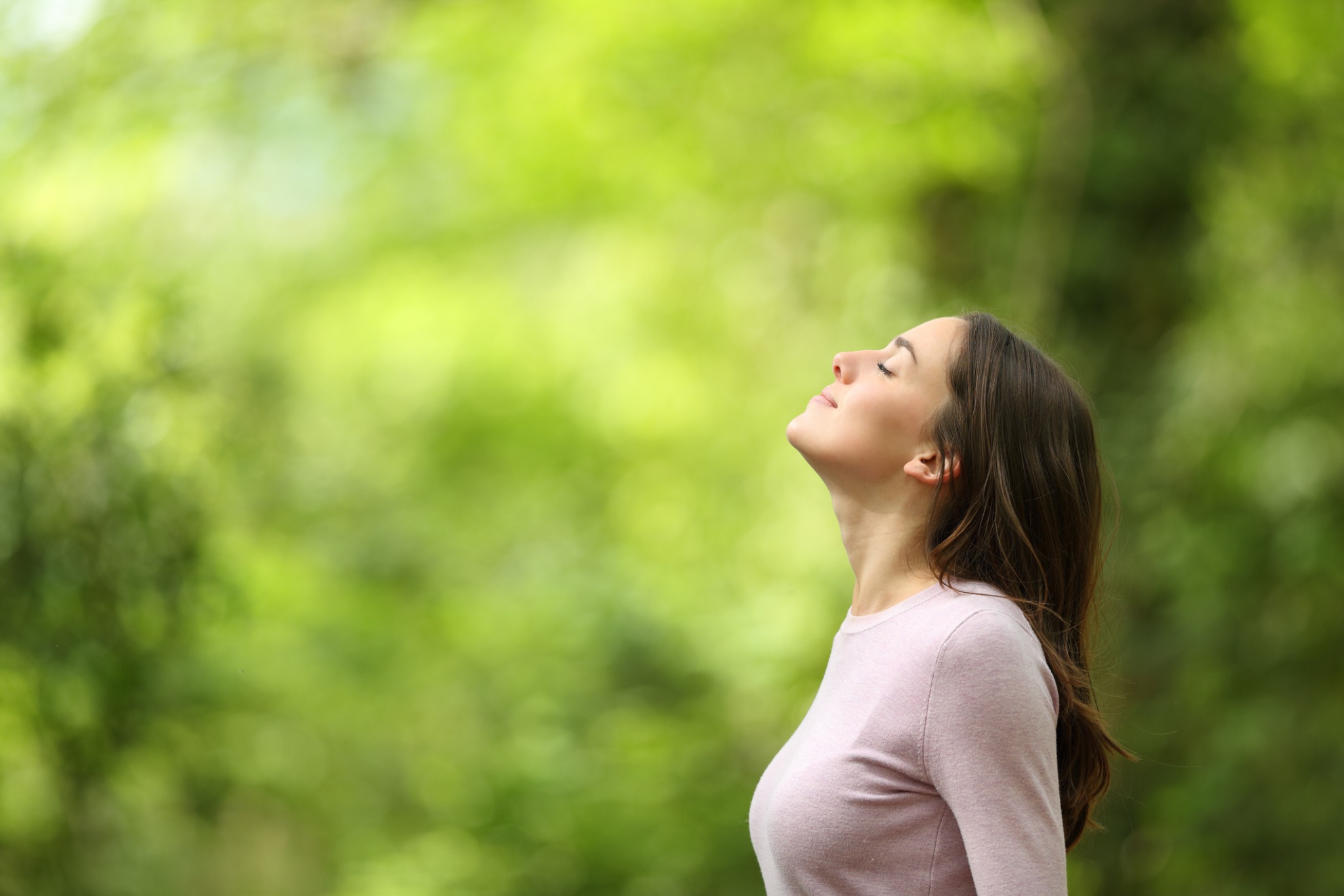 A woman outdoors with her eyes closed, surrounded by green foliage.