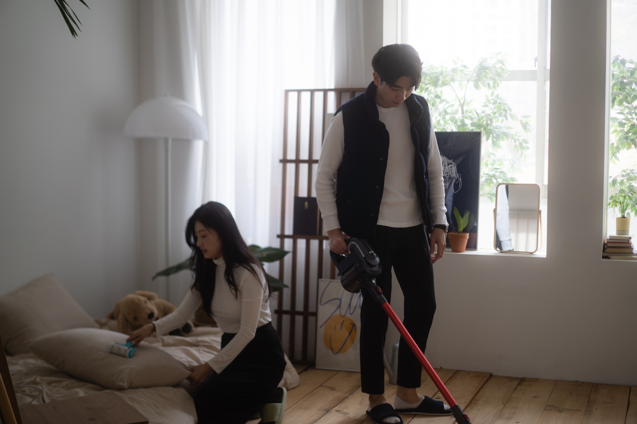 Two adults cleaning a room together, one vacuuming and the other using a lint roller.