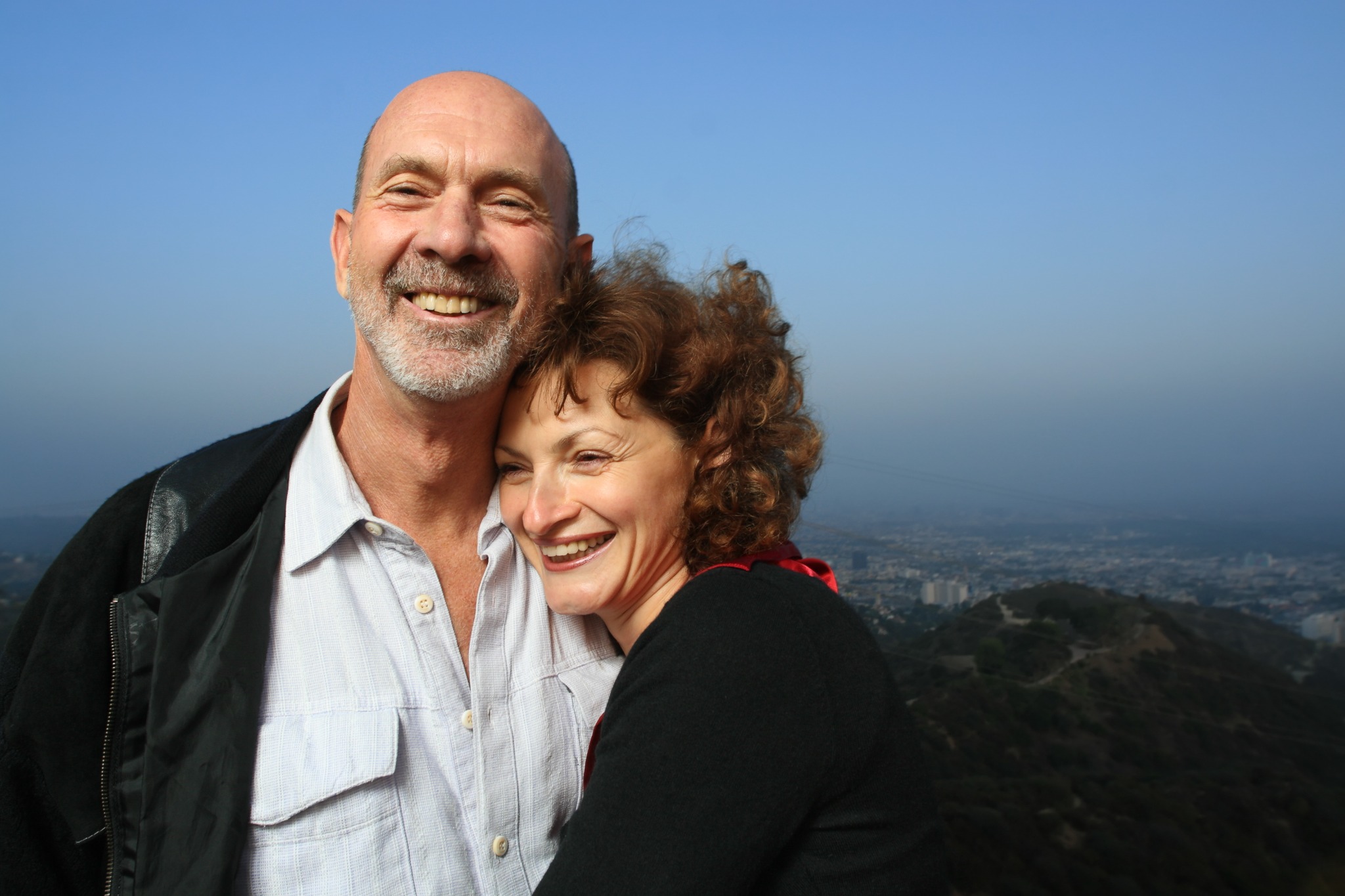 Two adults embracing outdoors with a city and hills behind them.