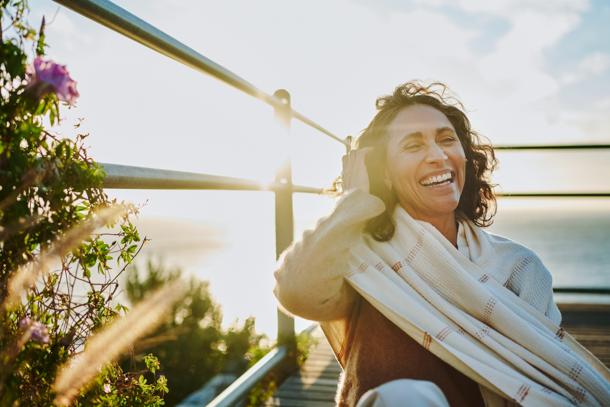 A woman laughing outdoors beside the ocean in warm evening light.