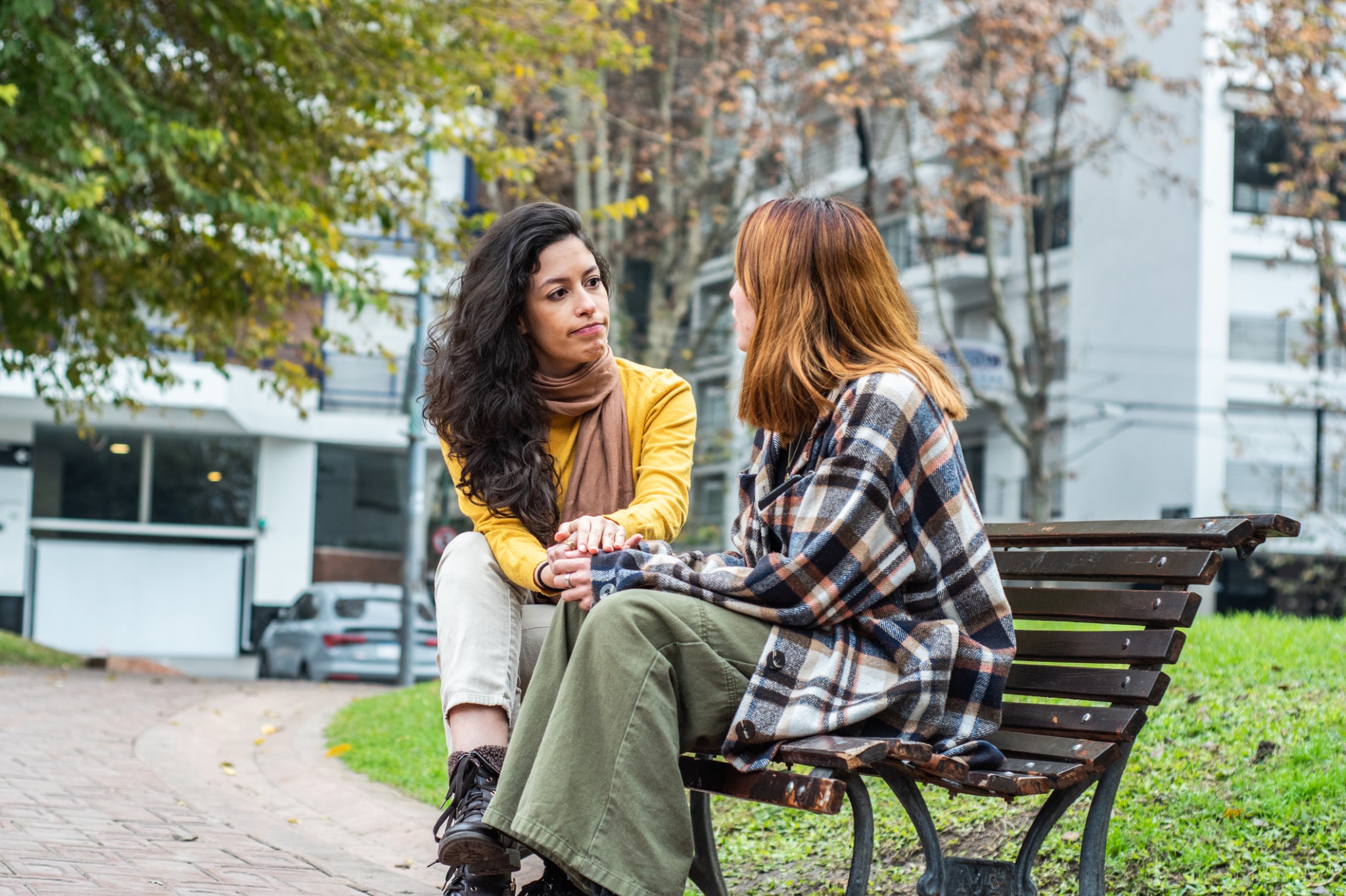 Two women sitting on an outdoor bench, one resting a hand on the other’s hands.