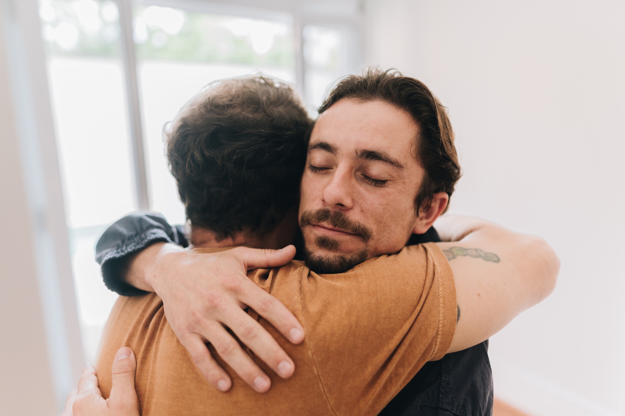 Two men embracing indoors, one with his eyes closed.