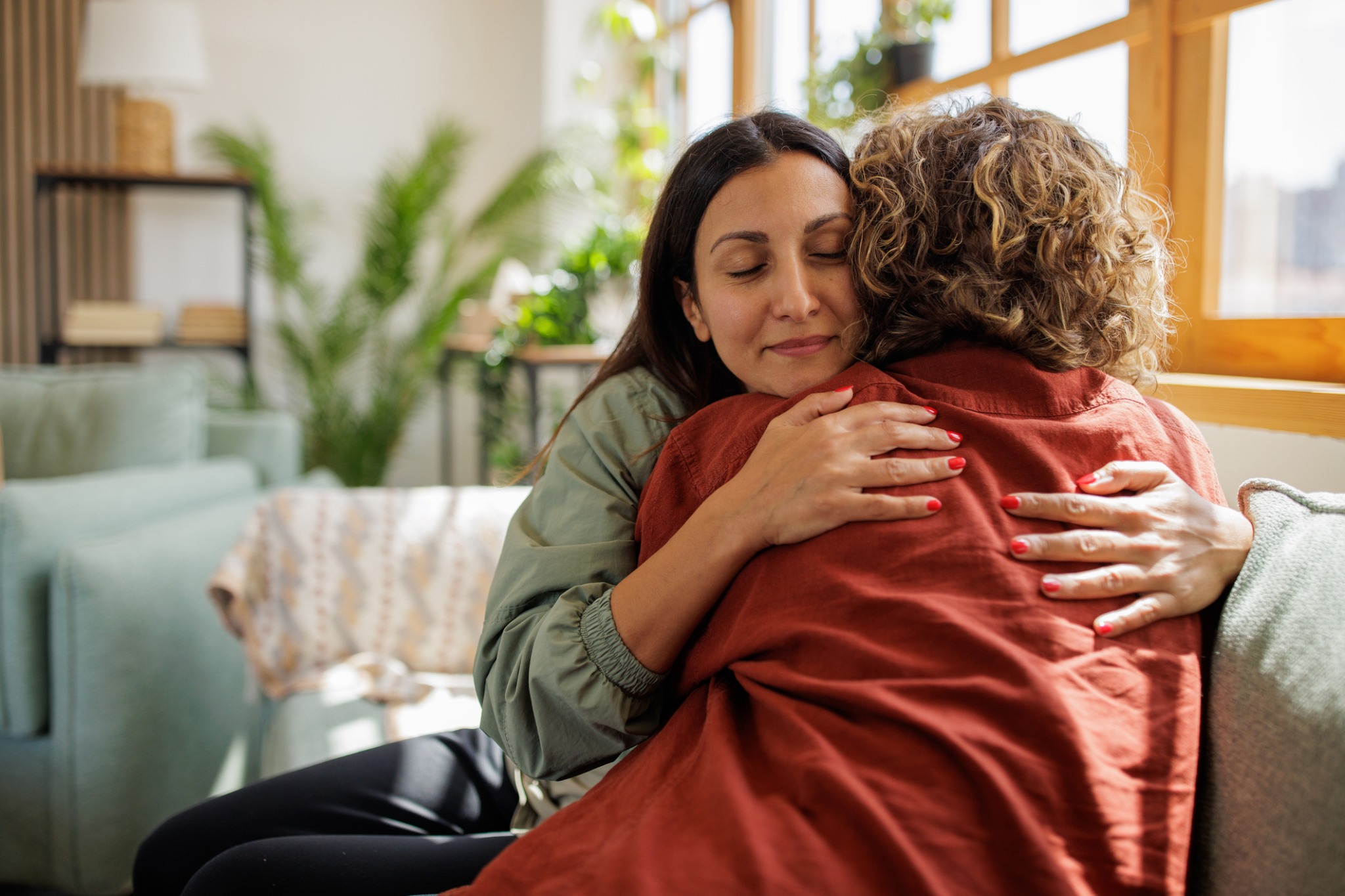 Two women embracing on a sofa in a sunlit room.