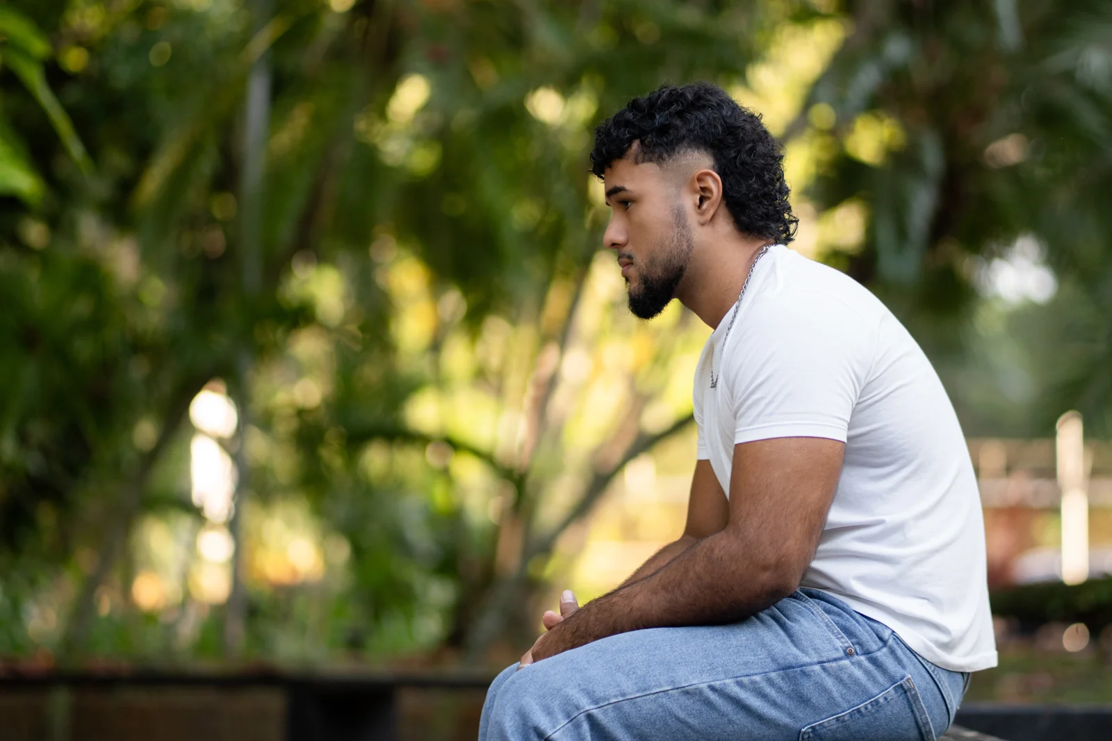 A man seated outdoors beside a leafy path, looking into the distance.