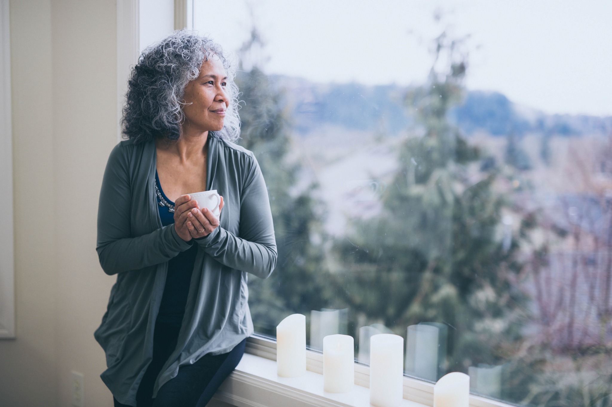A woman holding a cup and looking out through a large window.