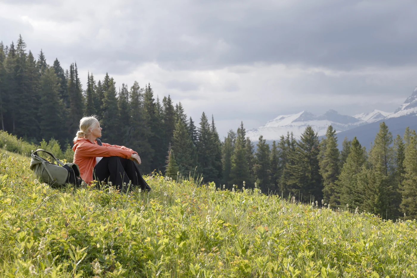 A person seated in a mountain meadow beside a backpack, looking toward the trees and mountains.